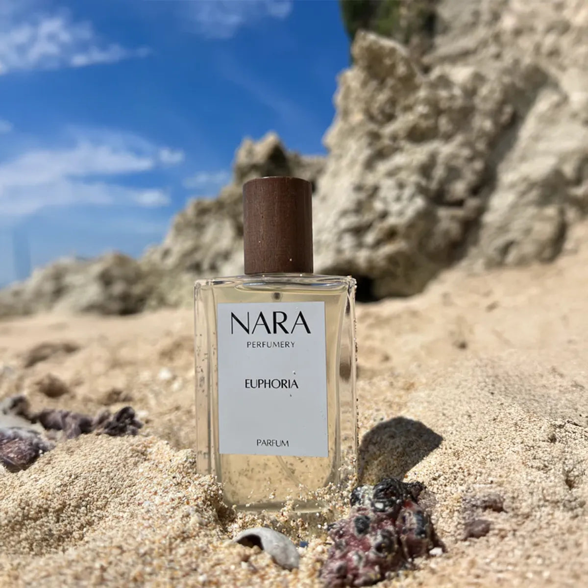 Nara Parfums Euphoria perfume bottle on a sandy beach with rocks and blue sky.