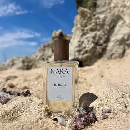 Nara Parfums Euphoria perfume bottle on a sandy beach with rocks and blue sky.