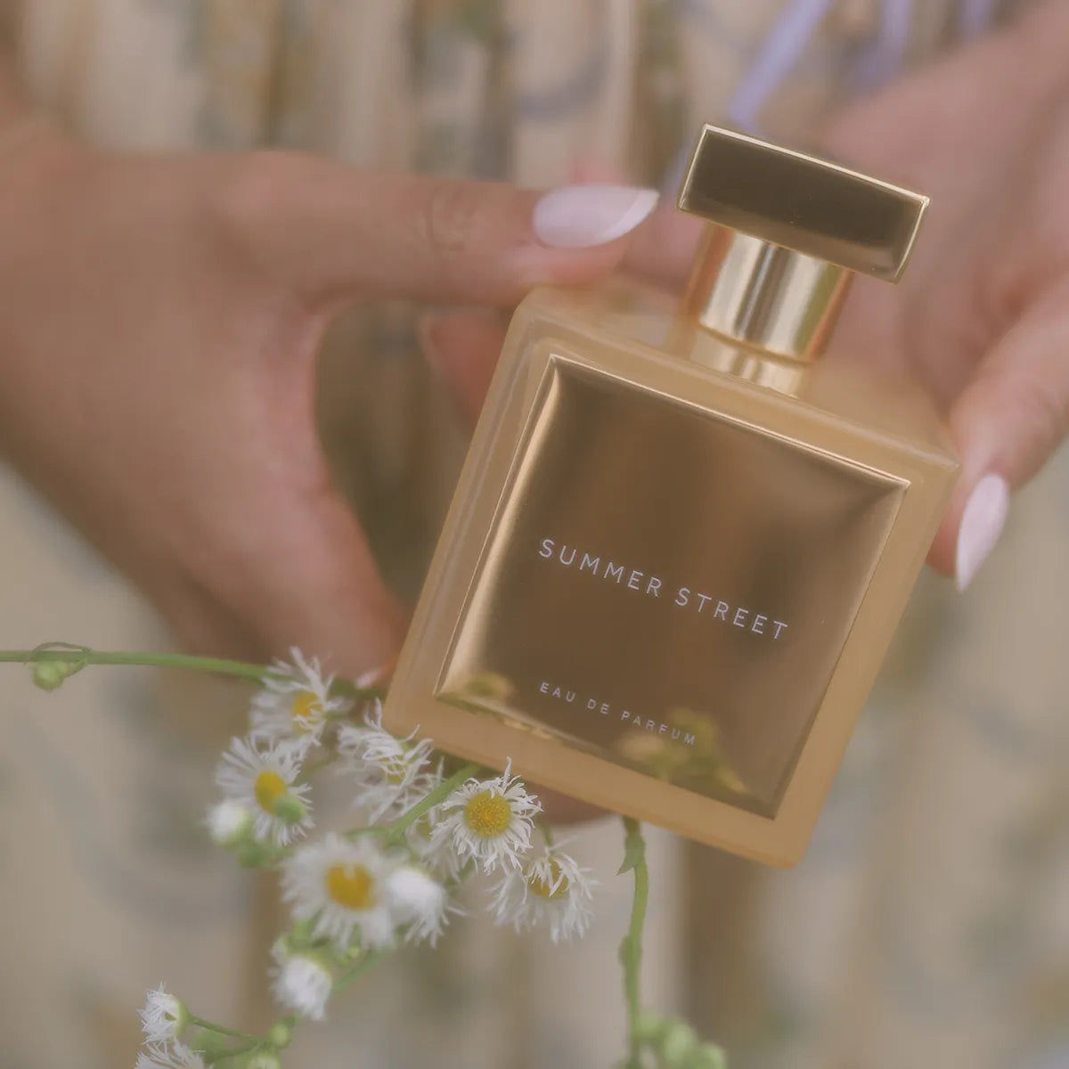 Gold 'Summer Street' perfume bottle held by a hand with flowers in the foreground