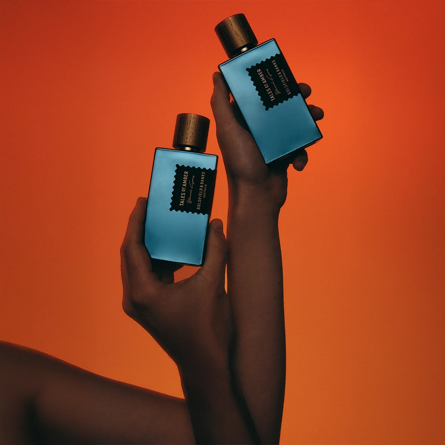 Two blue perfume bottles held by hands against an orange background