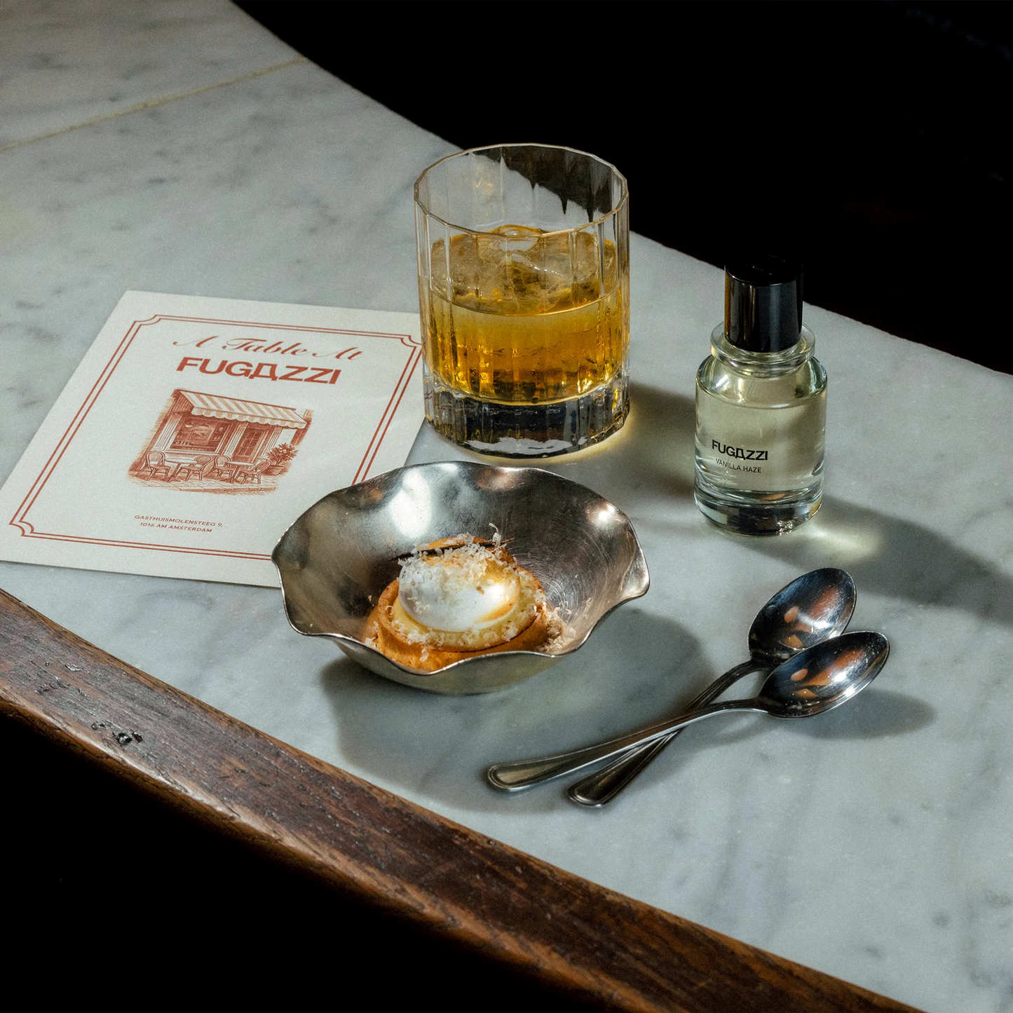 Glass of whiskey with ice, small dish, spoons, and Fugazzi bottle on a marble surface.